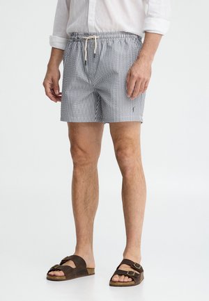 Hombre con camisa blanca de manga larga, pantalones cortos con cordón a rayas azules y blancas, y sandalias marrones con hebilla, de pie sobre fondo blanco.