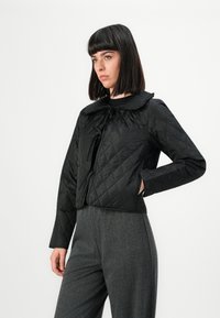 Jeune femme avec des cheveux noirs raides portant une veste matelassée noire avec un col volantré et un pantalon taille haute gris, se tenant avec une main sur la hanche.