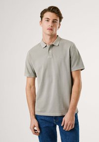 Polo shirt gris clair à manches courtes, col boutonné et logo discret ; associé à un jean en denim bleu. Tissu doux et texturé.
