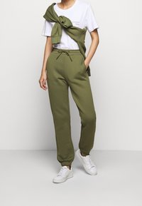 Grüne Jogginghose mit elastischen Bündchen und verstellbarem Kordelzugbund, kombiniert mit einem weißen T-Shirt. Leichter Pullover über die Schultern gelegt.