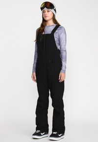 Salopettes noires avec bretelles réglables, dotées d'une fermeture éclair à l'avant, de poches latérales, portées sur un t-shirt à manches longues violet clair. Baskets noires.