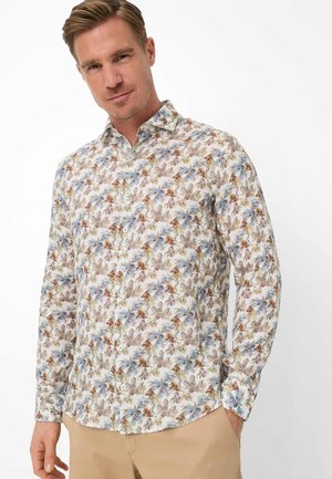 Mann trägt ein langärmliges Hemd mit blauen und braunen Blumenmustern, kombiniert mit beigen Hosen, und steht vor einem weißen Hintergrund.