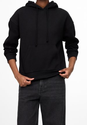 Personne portant un sweat à capuche noir avec poche avant et cordons, associé à un jean gris foncé, les mains ajustant l'ourlet du sweat.