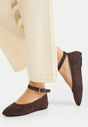 Braune Wildleder-Ballerinas mit spitzer Zehenpartie, Knöchelriemen mit Schnalle und flacher Sohle, kombiniert mit hellfarbenen, gekürzten Hosen.