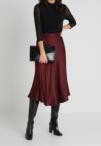 Haut noir transparent à manches longues avec motif en diamant, associé à une jupe midi en satin bordeaux et une pochette noire en relief croco. Bottes noires à talons hauts.