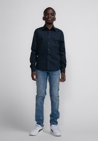 Navy blauwe button-up shirt met lange mouwen, gecombineerd met lichtblauwe jeans en witte sneakers met zwarte accenten. Glad materiaal en klassieke pasvorm.