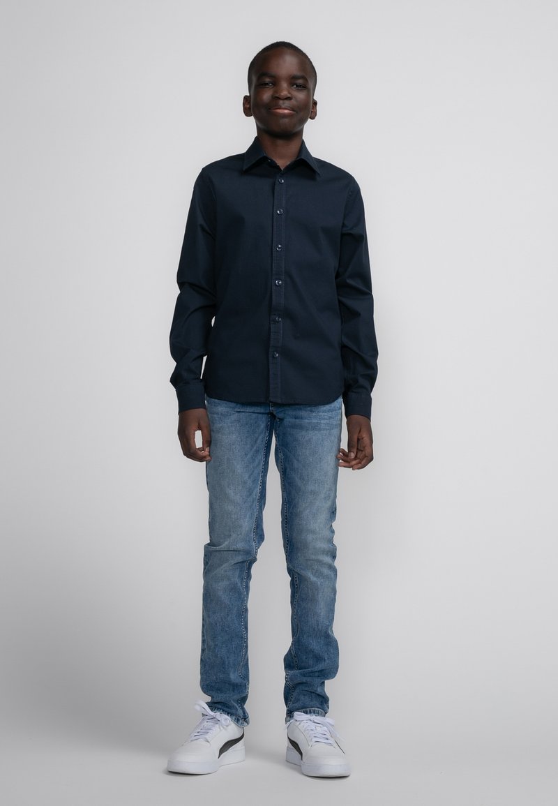 Navy blauwe button-up shirt met lange mouwen, gecombineerd met lichtblauwe jeans en witte sneakers met zwarte accenten. Glad materiaal en klassieke pasvorm.