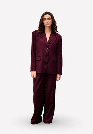 Vrouw die staat, gekleed in een loszittend donker bordeaux pak met brede broekspijpen en een dichtgeknoopt blazer, tegen een effen lichte achtergrond.
