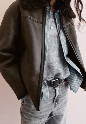 Personne portant une veste en peau retournée marron foncé sur un pull gris et une chemise rayée, avec un jean délavé clair et une ceinture noire, les mains dans les poches.