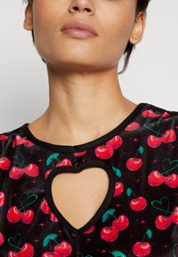 Vestido de veludo preto com um recorte em forma de coração na gola e um vibrante padrão de cerejas em vermelho e verde. Detalhes em preto.