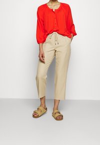 Rød, langærmet, knappet bluse med en samlet hals; beige, løstsiddende, cropped bukser med elastisk snor; brune sandaler.