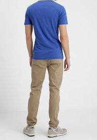 Mężczyzna stojący tyłem, ubrany w niebieską koszulkę, beżowe spodnie dresowe i szare sneakersy New Balance na białym tle.