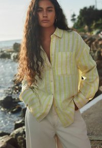 Camicia button-up giallo chiaro con righe verdi, dotata di tasca frontale e vestibilità rilassata, indossata sopra pantaloni color crema. Texture del tessuto naturale.