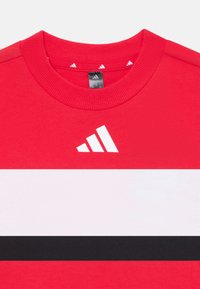 Red t-shirt featuring a white and black horizontal stripe design, with a prominent white Adidas logo on the chest.