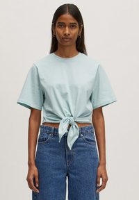 Helles, blaues, kurzärmliges Cropped-T-Shirt, vorne geknotet, kombiniert mit hoch taillierten blauen Jeans. Glattes Baumwollmaterial.