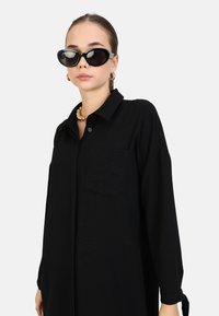 Zwarte jurk met knopen aan de voorkant, lange mouwen, een borstzak en een zachte textuur. Het model draagt oversized zwarte zonnebril.