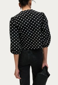 Blouse noire à pois blancs, manches bouffantes et col arrondi. Associée à un jean noir, tenant une pochette noire texturée.