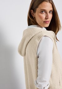 Gilet en polaire couleur crème avec capuche, présentant une surface texturée douce. Porté par-dessus une chemise blanche à col, mettant en valeur le design en couches.