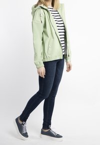 Kvinna klädd i en ljusgrön hoodie, svart- och vitrandig tröja, mörka slim-fit-jeans och marinblå sneakers, ståendes på sidan.