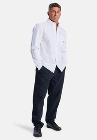 Weißes Hemd mit Kragen und Brusttasche, kombiniert mit dunkeln marineblauen Hosen und schwarzen Sneakern für ein kontrastierendes Outfit.