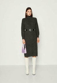 Egy nő sötétszürke kötött ruhában, fekete övvel, egy kis levendula színű táskával és fehér térdcsizma mellett, egy sima háttér előtt.