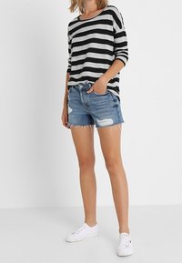 Czarna i szaro-skalista bluzka z długim rękawem w połączeniu z podartymi szortami dżinsowymi. Modelka nosi białe tenisówki, co podkreśla swobodny i luźny fason.