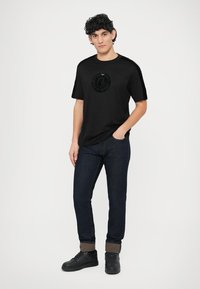 Melna īss piedurkņu T-krekls ar apaļu logotipu priekšpusē, kopā ar tumši ziliem džinsa džinsiem un melniem sporta apaviem.