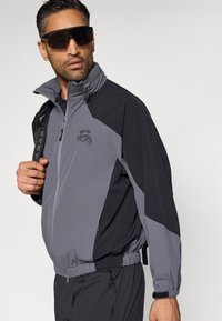 Γκρι και μαύρο αδιάβροχο μπουφάν με φερμουάρ, ψηλό γιακά, ελαστικούς μανσέτες και λογότυπο. Διαθέτει πάνελ σε διαφορετικά χρώματα και άνετη εφαρμογή.