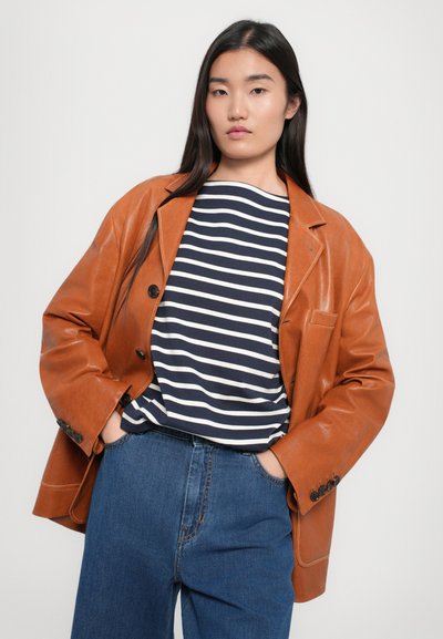 Blazer in pelle marrone con rever a punta, camicia a righe scure sotto, e jeans in denim blu, con bottoni sui polsini e una vestibilità casual.