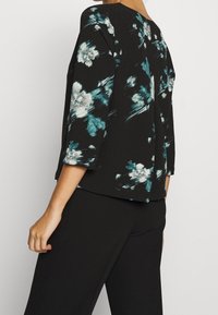 Sort blomsterbluse med teal og hvide mønstre, rund halsudskæring, trekvartærmer og løs pasform, parret med sorte bukser.