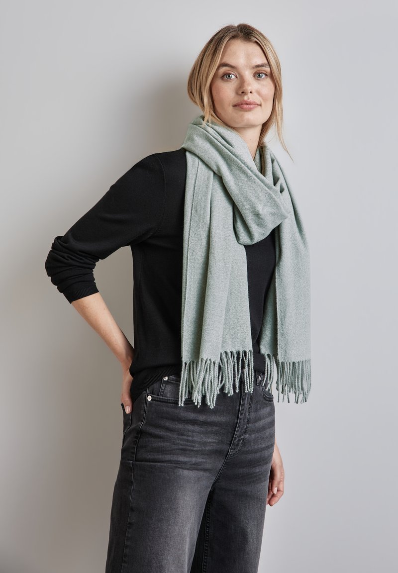 Lys grøn tørklæde lavet af blødt stof, med frynset kant. Båret løst omkring halsen, komplementeret af en sort top og grå jeans.