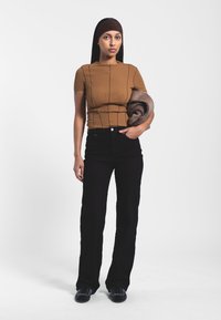 Top marrón de manga corta de canalé, jeans negros de cintura alta y acampanados, sosteniendo una prenda marrón enrollada. Colores sólidos, silueta ajustada, textura suave.