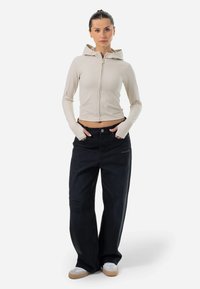 Světle béžová mikina na zip s přiléhavým střihem a kapucí, spárovaná s volnějšími černými džíny z denimu se zvýrazněným opotřebením.
