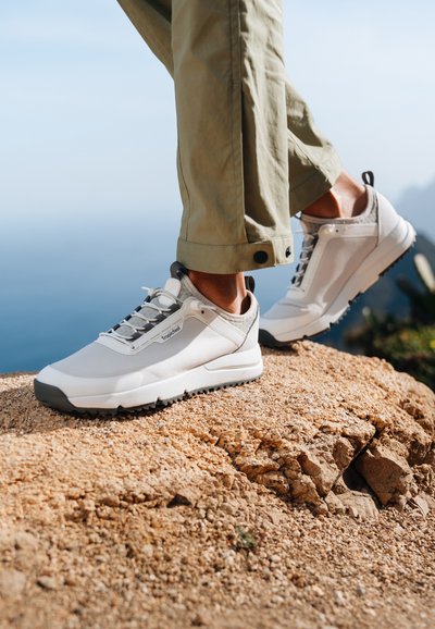 Scarpe da esterno bianche con dettagli grigi, tomaia in mesh texturizzato e suole robuste, posizionate su un terreno roccioso con uno sfondo panoramico.