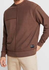Uomo che indossa una felpa a maniche lunghe in patchwork marrone con un'etichetta nera vicino all'orlo e pantaloni beige, con la mano appoggiata sui fianchi.