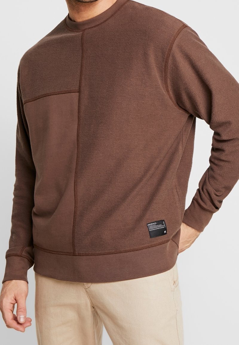 Uomo che indossa una felpa a maniche lunghe in patchwork marrone con un'etichetta nera vicino all'orlo e pantaloni beige, con la mano appoggiata sui fianchi.