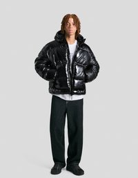Shiny black puffer jacket with a zipper, hood, and wide quilted design; paired with loose dark jeans and chunky black sneakers.