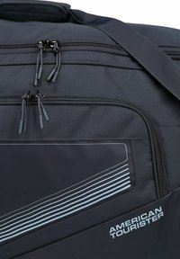 Sac de voyage en tissu gris avec des accents noirs, doté de deux zippers, d'un logo et de rayures diagonales bleu clair sur le côté.