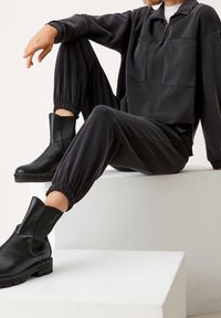 Felpa nera con zip e due tasche frontali, abbinata a pantaloni joggers coordinati con polsini elasticizzati. Stivali neri alla caviglia con finitura testurizzata.