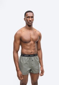 Μυώδης άνδρας με κοντά μαλλιά, ντυμένος με γκρι μποξέρ σορτς με μαύρη μέση που φέρει την ετικέτα "HUGO", στέκεται μπροστά σε απλό φωτεινό φόντο.