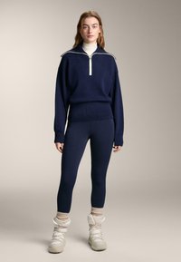 Marineblauer Strickpullover mit Reißverschlusskragen, enganliegende Leggings und weiße Schneestiefel. Der Pullover hat einen gerippten Saum und kontrastierende Kragen-Details.