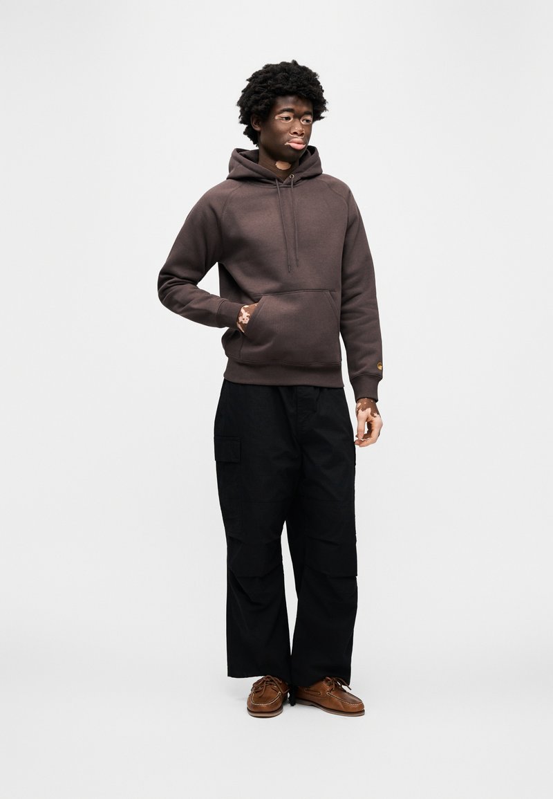 Brun hættetrøje med en forlommelommer, båret med sorte cargobukser. Modellen står på en ensfarvet baggrund iført mørkebrune sko.