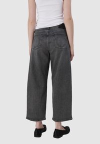 Szerokie, szare dżinsowe spodnie cechują się wysokim stanem, dwoma tylnymi kieszeniami z ozdobnym przeszyciem oraz surowym wykończeniem u kostek.