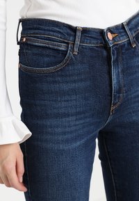 Tmavě modré džíny z denimu s viditelným šitím, pětibetovým designem a měděnými doplňky. Mají slim fit a jemnou texturu.