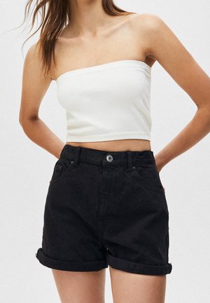 Frau trägt ärmelloses weißes Crop-Top und hochgekrempelte schwarze Denim-Shorts, steht mit den Händen hinter dem Rücken vor schlichtem hellem Hintergrund.