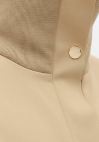 Top beige con una textura suave y un cuello acanalado, complementado con un detalle de botón dorado brillante en el lateral.