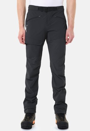 Mann trägt schwarze Outdoor-Hose mit Reißverschlusstaschen, einer Klappentasche am Oberschenkel und Wanderschuhe mit orangefarbenen Sohlen.