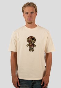 Beige Baumwoll-T-Shirt mit einem zentralen gestickten Design einer Puppe mit einem Totenschädel, floralen Details und einem schwarzen Umriss. Rundhalsausschnitt, kurze Ärmel.