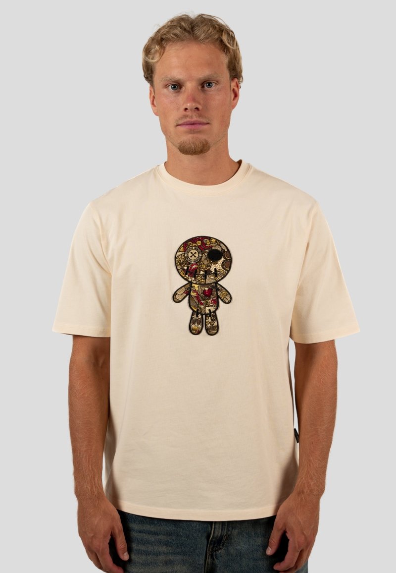 Beige Baumwoll-T-Shirt mit einem zentralen gestickten Design einer Puppe mit einem Totenschädel, floralen Details und einem schwarzen Umriss. Rundhalsausschnitt, kurze Ärmel.