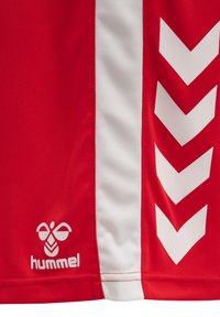 Camiseta deportiva roja con franjas laterales blancas. Presenta un patrón en forma de chevron en blanco y un logo de "hummel" en blanco en la parte inferior. Textura de tela suave.